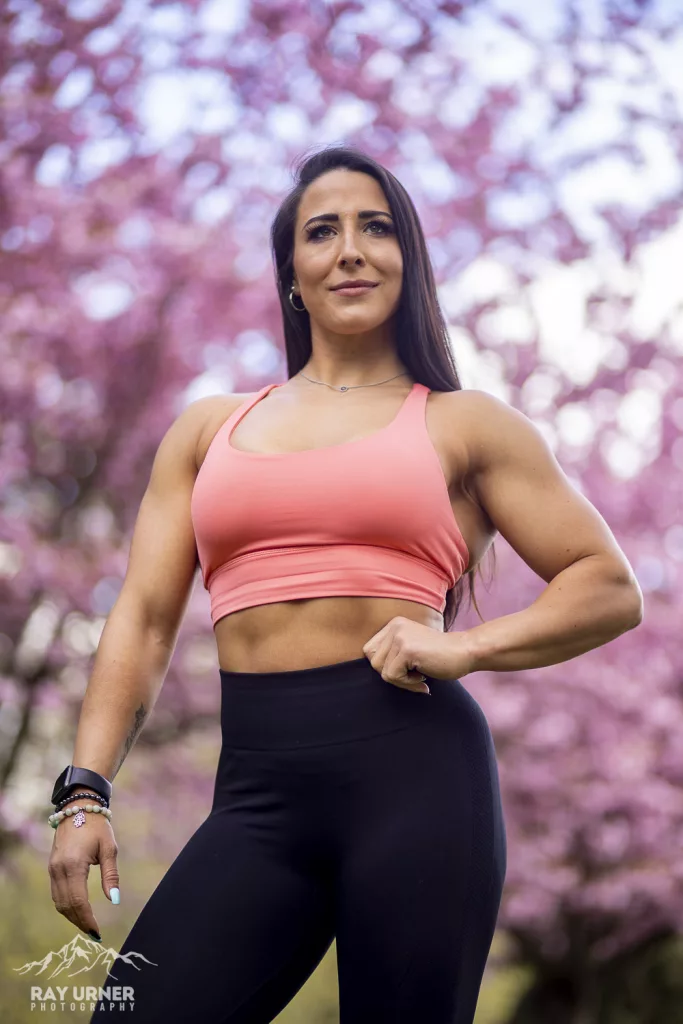 Figure competitor standing in a park with cherry blossom trees behind her